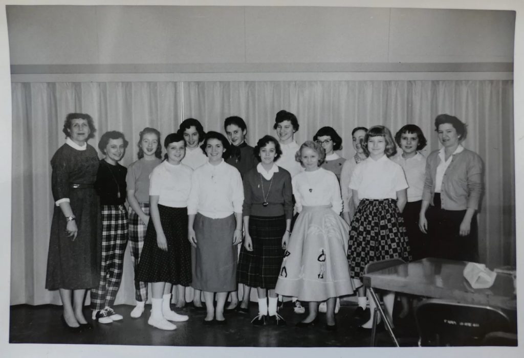 1950s vintage photo of a group of girls posing for their school club photo. The girls are wearing poodle skirts / circle skirts, saddle shoes and other 1950s fashions. 