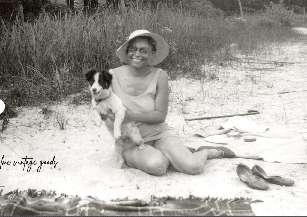 1920s snapshot of a Black woman seated on a blanket in the sand holding a cocker spaniel while wearing a floppy hat, swimsuit and shoes.