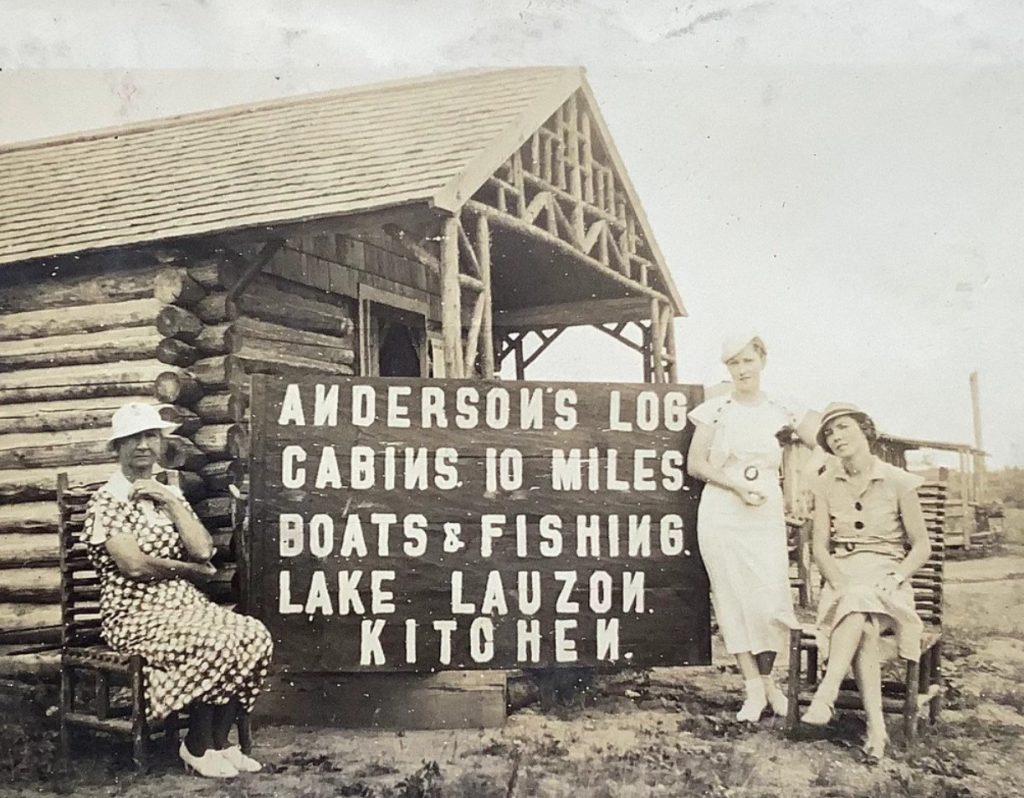 1930s vintage photo of women in 1930s dresses posing in front of a sign advertising log cabins to rent and boats and fishing. 