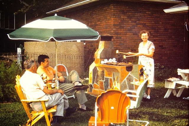 1950s / 1960s vintage photo of a family in their backyard cooking on an outdoor stove and sitting in various outdoor chairs. 