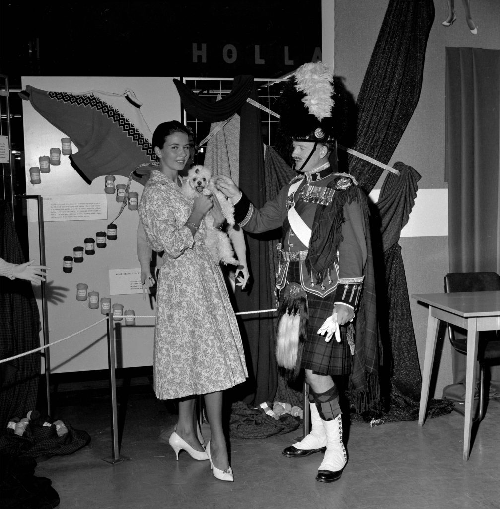 1950s vintage photo of the 1959 Australia exhibit featuring an image of a woman in a 1950s dress holding a dog with a bagpiper 