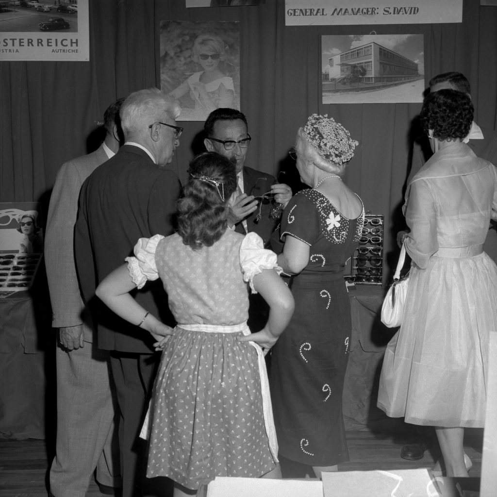 1960s vintage photo of a woman in a Dirndl at the Austria Exhibit at the Toronto CNE in 1960. She is surrounded by others in early 1960s fashions. 