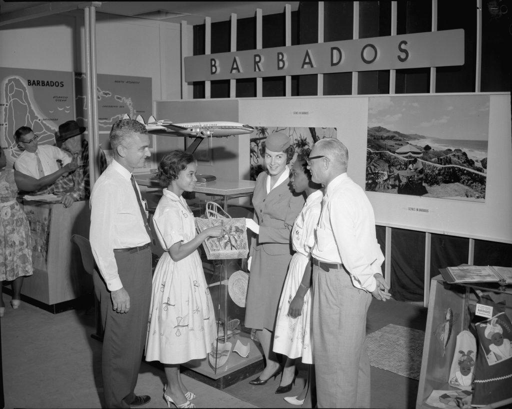 1960s vintage photo of the The Barbados T.C.A. exhibit in 1960 featuring all the places you can go with the airline. The image features a TCA flight attendant and two young Black girls in pretty 1960s dresses and a fantastic 1960s straw purse. 