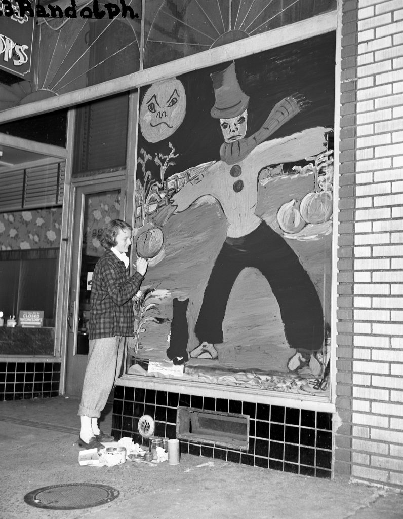 1940s vintage image of a teenage art student in 1948 painting a Halloween scene on a store front window featuring a black cat and a scarecrow. 