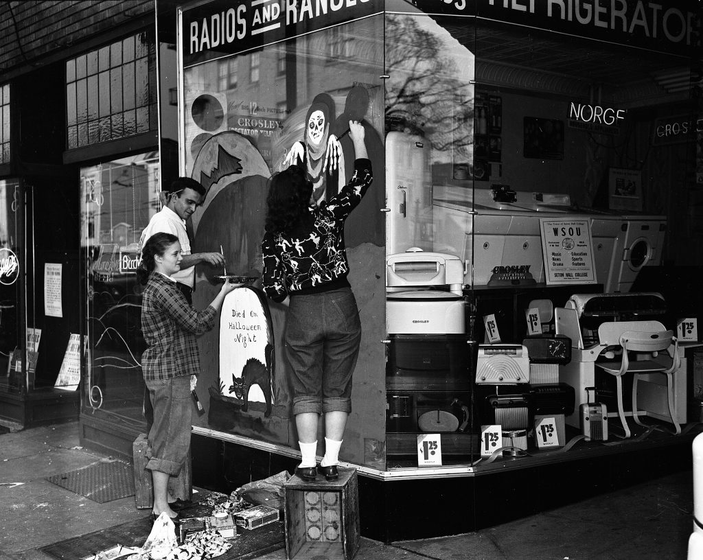 1940s vintage image of art students in 1948 painting a Halloween scene on a store front window selling radios, ranges and other items. The teenagers are wearing 1940s fashions for Teens / Juniors. One woman is wearing a swing dance / Lindy Hop sweater. 