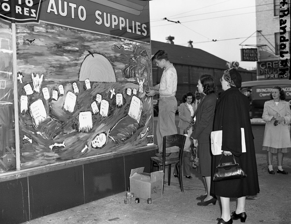 1940s vintage image of a boy in Newark, NJ painting a Halloween mural on a auto supplies store of a graveyard scene. 