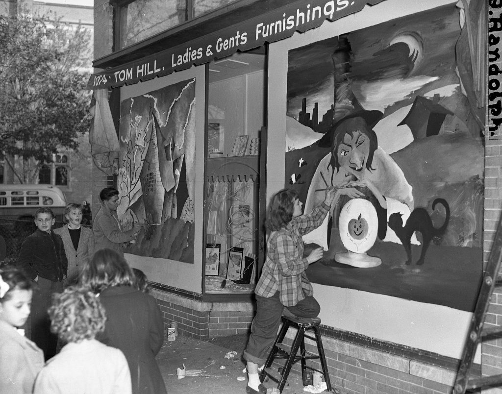 1940s vintage image of teenagers in Newark, NJ painting a Halloween mural on a furniture store. The mural is of a witch with a black cat and a haunted house. 