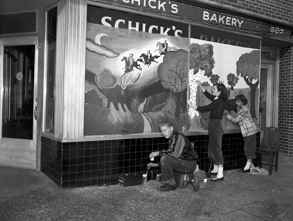 1940s vintage image of a teenage art student in 1948 painting a Halloween scene on a store for a bakery in Newark, NJ. The Halloween mural features skeletons riding horses in the sky. 