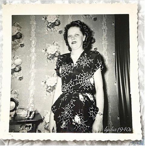 1940s vintage photo of a woman in a 1940s dress with a spider web floral pattern on it. She is standing in front of a floral wallpaper. 