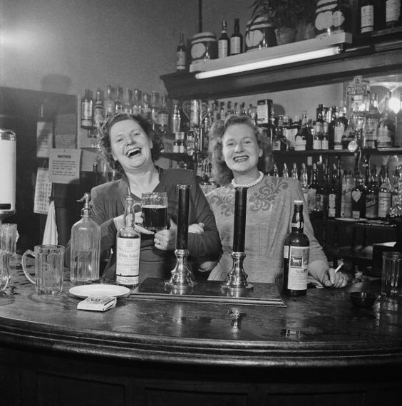 1940s vintage photo of Two barmaids at a pub in London, 1949 in 1940s dresses and 1940s hairstyles holding beers and cigarettes. 