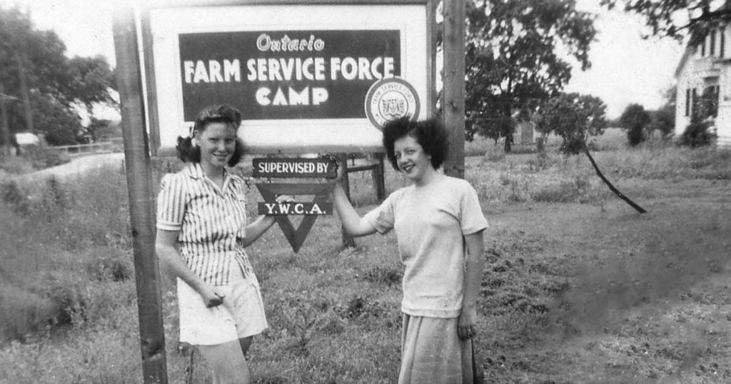 1940s vintage photo of two young women in 1940s fashion posing in front of a Sign that says "Ontario Farm Service Force Camp". They were Farmerettes and they were farming for the war effort at home in Canada. 