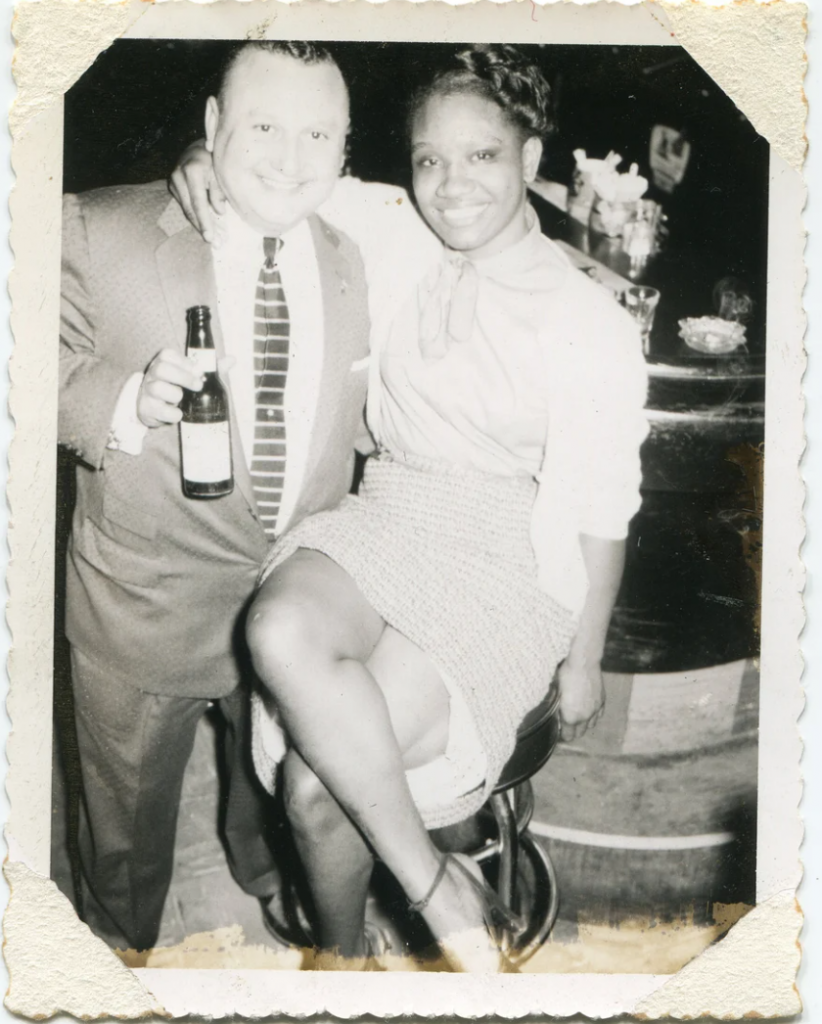 1950s vintage photo of a pretty Black woman in 1950s fashion sitting on a bar stool posing with a man in a 1950s suit holding a beer