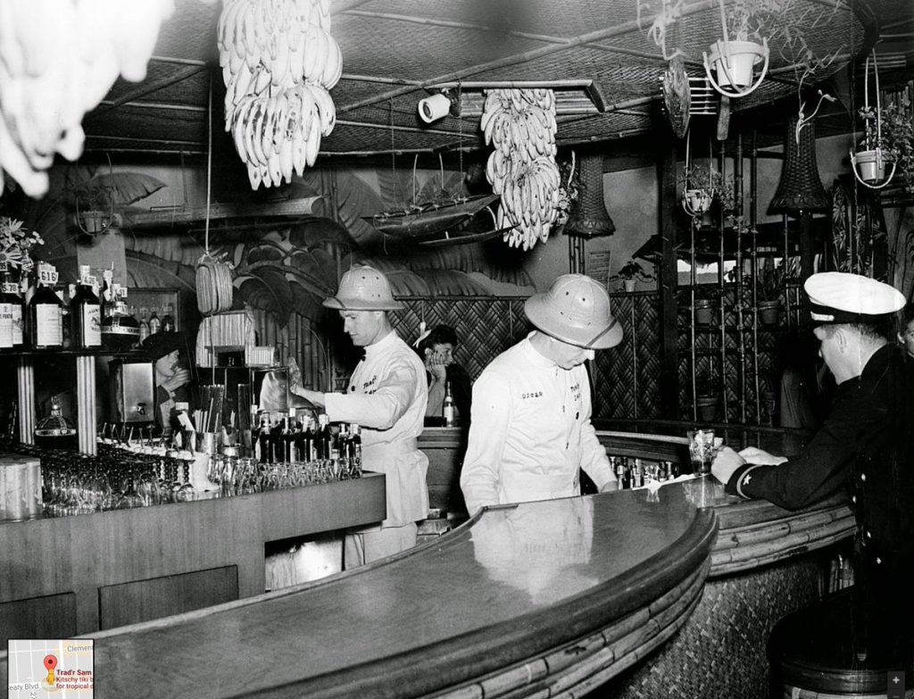 1950s vintage photo of bartenders working at the Tiki bar Tradr Sam they are wearing safari outfits a man in uniform sits at the bar a woman in the back enjoys a beer