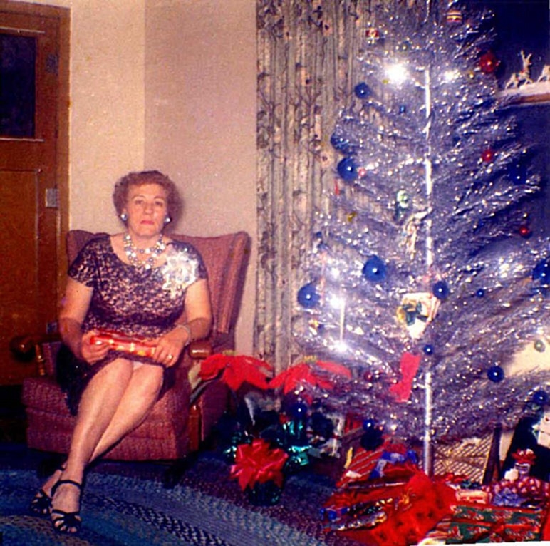 1960s vintage photo of a well dressed woman in a cocktail dress holiding a present and posing beside her Aluminum Christmas Tree.