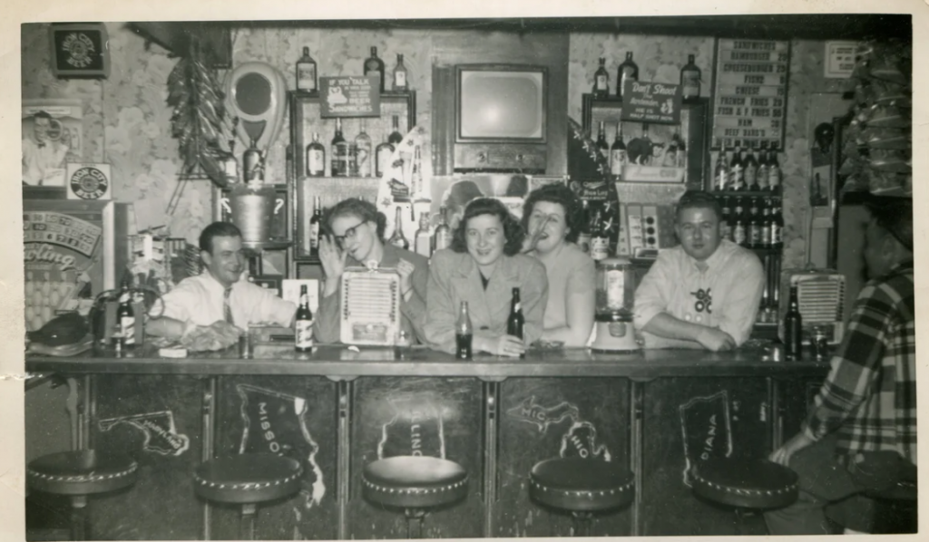 late 1940s /early1950s vintage photo of a group of young people behind a very decked out bar with the bartender tp the left. They look like they could be college students. 