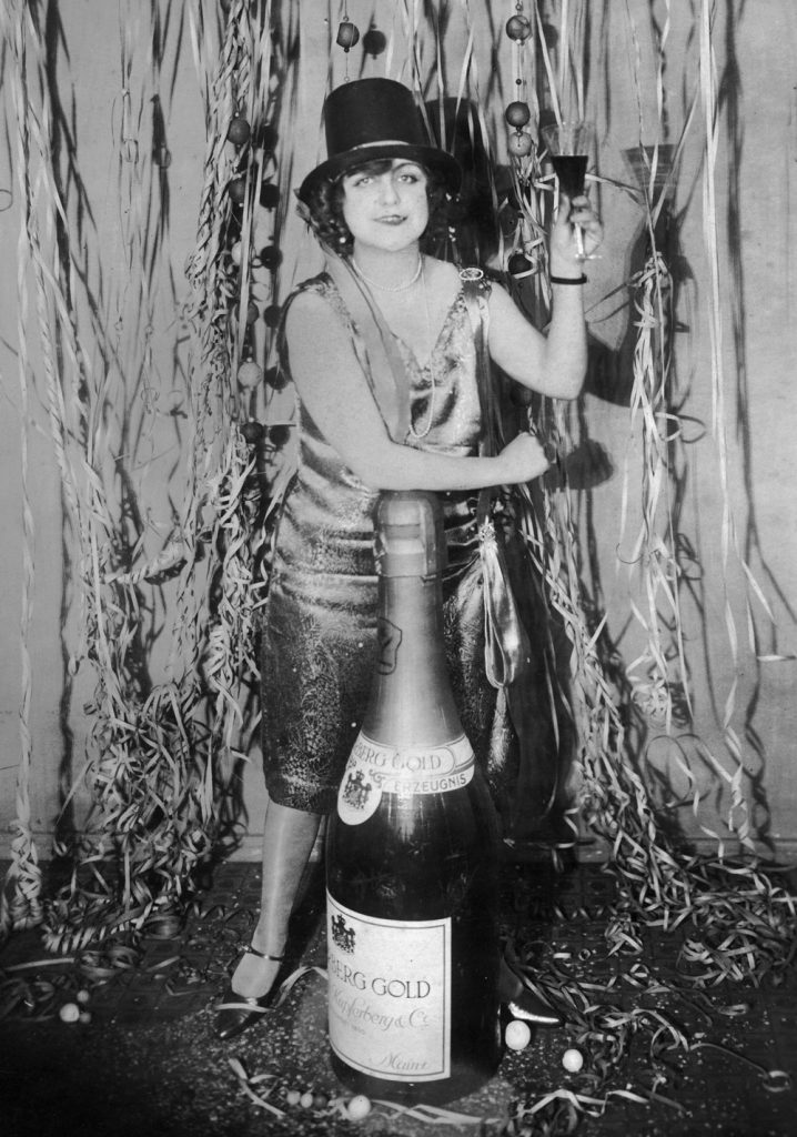 1920s vintage photo from a New Years Eve Party. A woman at a New Year's Eve party in Germany leans on an oversized bottle of champagne while making a toast, circa 1925.