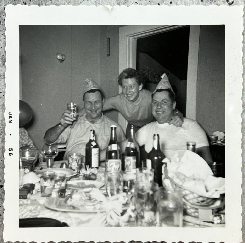 1950s vintage photo of a New Years eve house party 1956. Photo is a group sitting around a table posing with hats and drinks and food in front of them