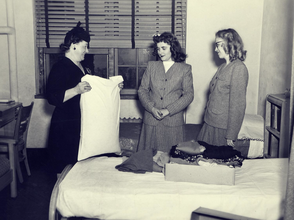 1940s Vintage photo of student life in a dorm at UW-Madison. An older woman holds a pillow while two younger women stand by an open suitcase in a dorm room. C. 1940s