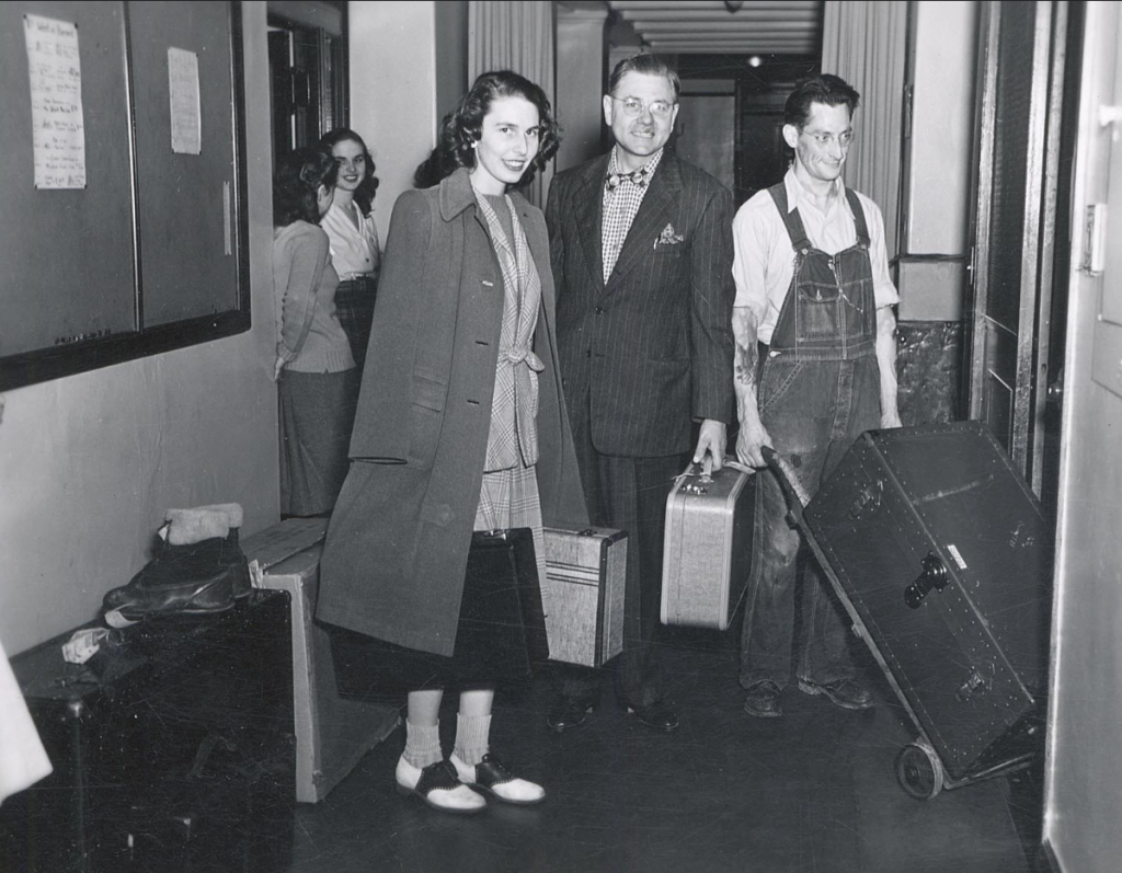 Early 1950s photo of Moving into the dorm; UW-Madison. A young woman holding luggage, accompanied by two older men also holding luggage, stands in the hallway of a dormitory. C. 1950s

 