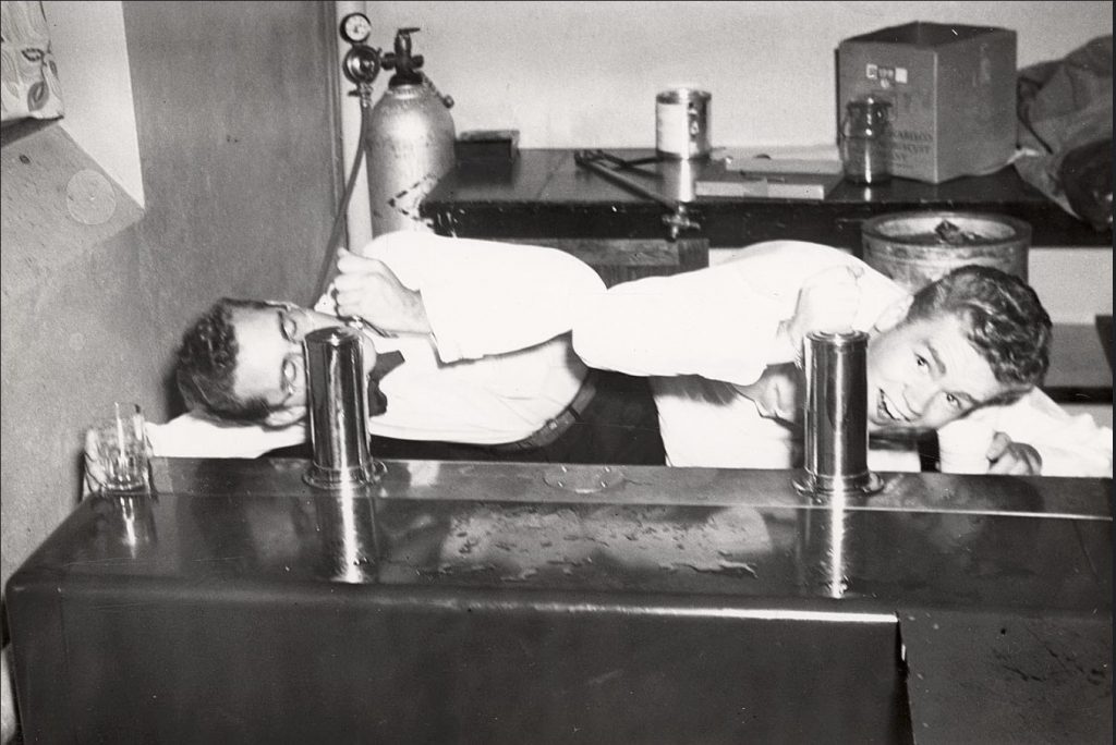 1940s vintage photo from the University of Wisconsin-Madison of 2 young men,  Jack Vazina and Dave Thompson, in waiter uniforms, drink from the taps at the bar during a party in Swenson house of Kronshage hall. C. 1949. Vintage Dorm Life. 