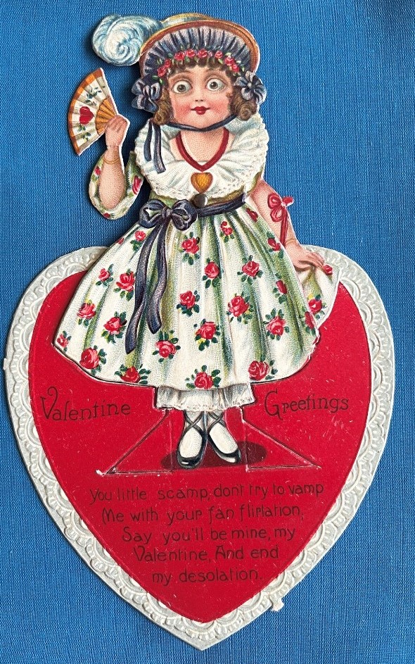 Vintage 3D moveable Valentine's card, 1920s. It's a pop-out, moveable card, the girl on the card is cut out so that she can stand up on the folded base, and her arms are hinged to move up and down. One hand holds a fan, the other holds up her dress with 'Valentine's Greetings' on a small heart hanging over her arm.