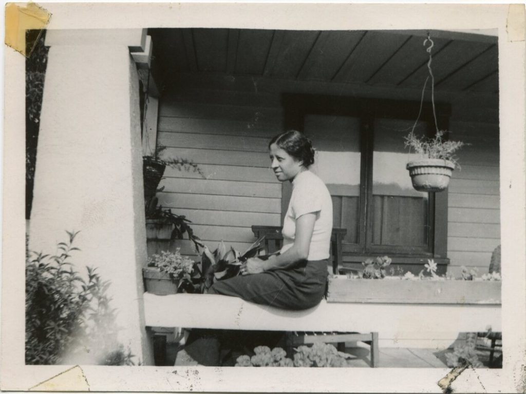 1930s vintage photo of a young Black woman in 1930s top and skirt posing on her front porch with all her flowers on her porch