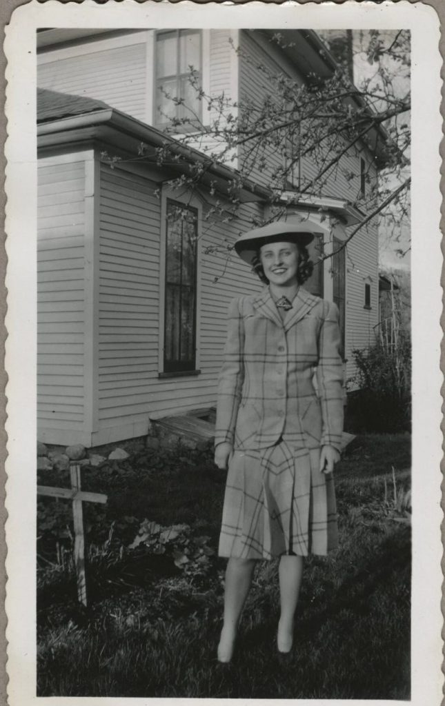 1940s vintage photo of a woman in a 1940s two piece suit in plaid and a 1940s hat with 1940s hairstyle posing in front of her house. Stunning example of 1940s fashions