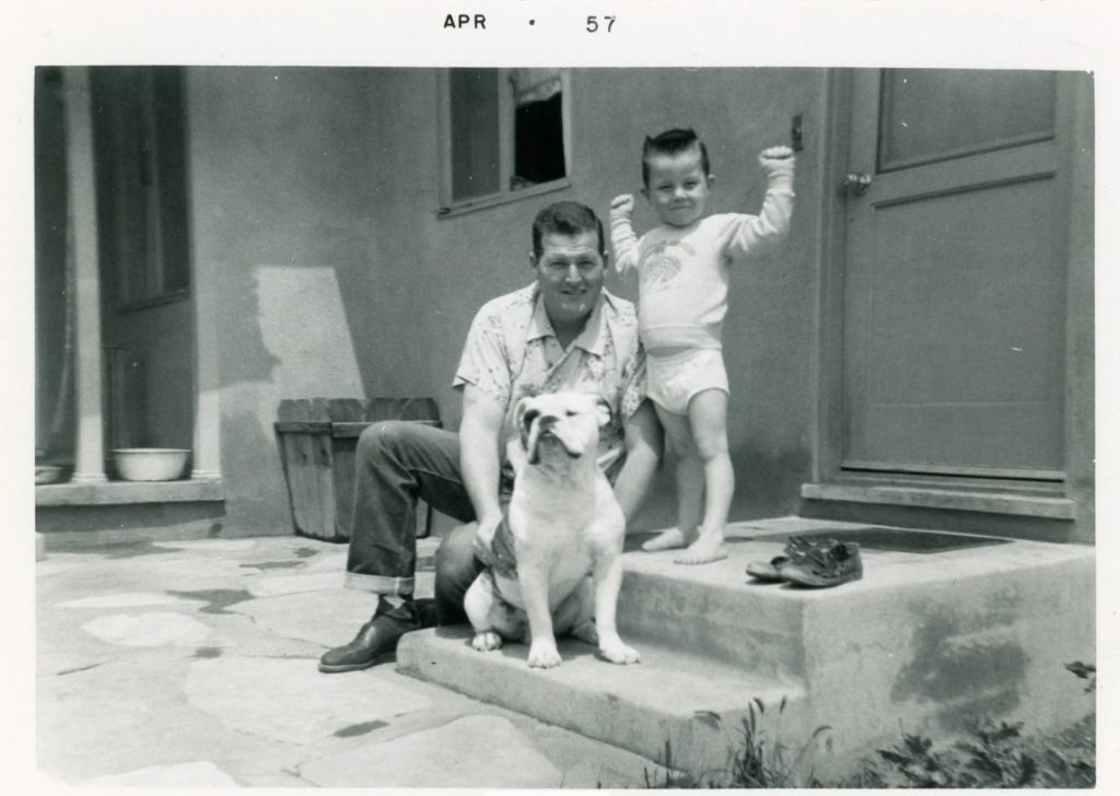 1950s vintage photo from april 1957 of a young child a toddler posing for a photo while flexing with his dad and his dog on the steps of their house