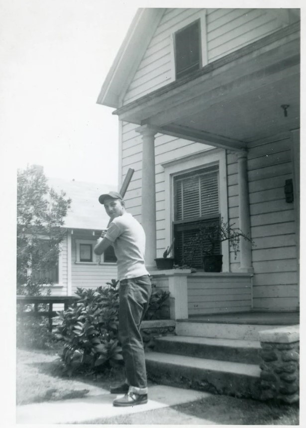 1950s vintage photo of a young man holding a baseball bat posing for a photo in front of his house