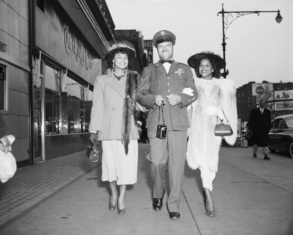 1950s vintage photo of two stylish Black women in 1950s fashions walking down the street during easter with a Black man in military uniform. 