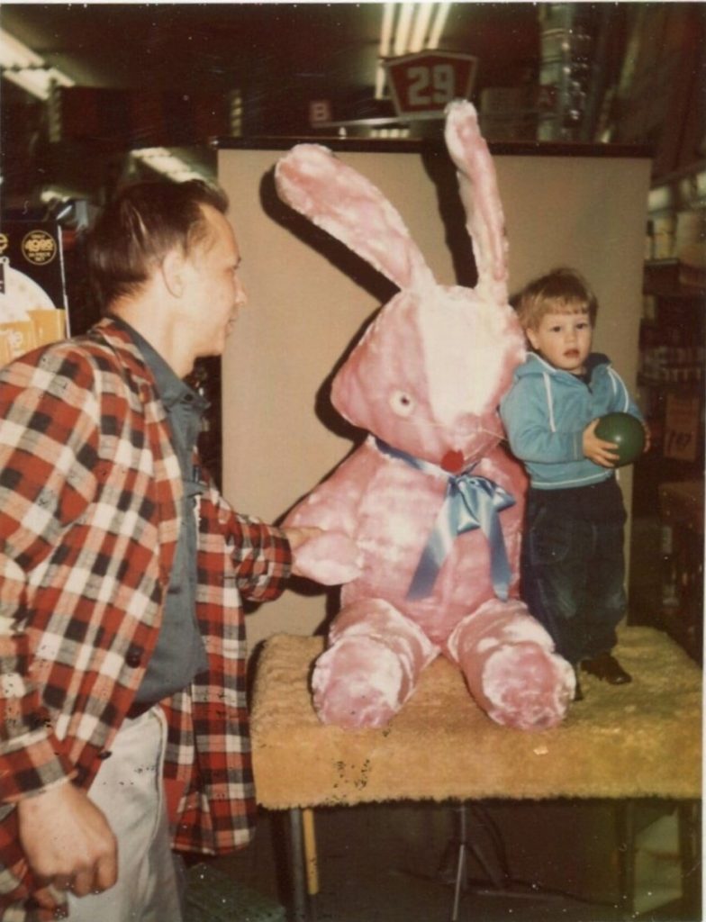 1960s photo of a young boy posing for a photo with a pink stuffed easter bunny while his dad ina flannel jacket holds up the toy