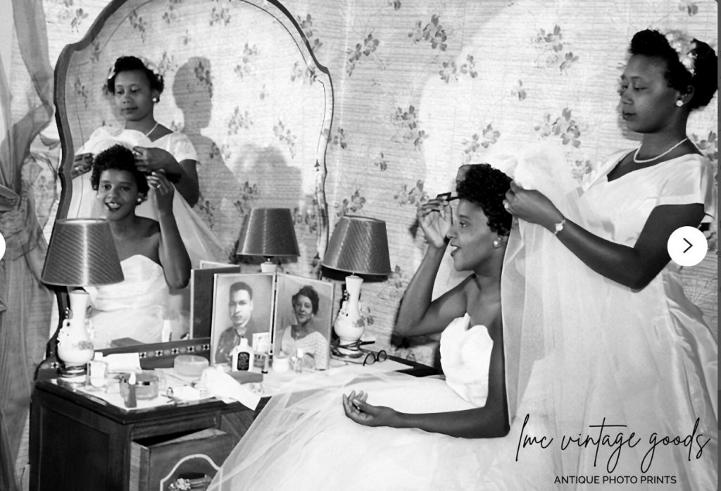 1950s Vintage Wedding Photo of a young Black Bride getting ready for her wedding day with her bridesmaid in her bedroom with a picture of her and her groom on the dressing table. 