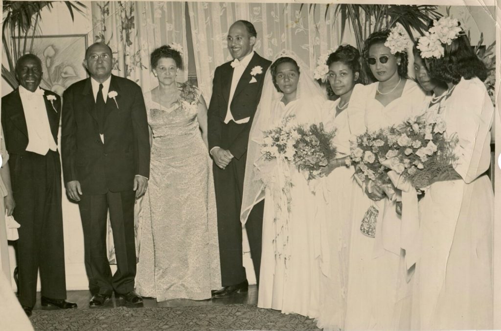 1940s Vintage Wedding photo of an African American Wedding party in their wedding outfits. The Black bride is stunning in her long veil and wedding gown and her Black groom is in a tuxedo. The Bridesmaids have beautiful hair flowers in their hair and large flower bouquets.