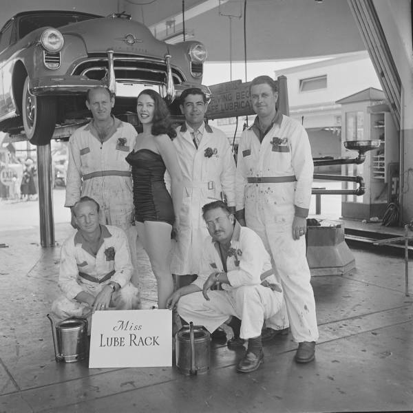 1950s vintage photo from 1951 of a beauty pageant featuring a woman in a 1950s swimsuit represening "Miss Lube Rack" posing in a garage with a car and the mechanics of the Muller bros dealership.