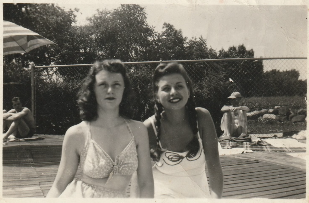 1940s vintage photo of two young women in 1940s swimsuits posing together at the pool. They both have 1940s hairstyles, one girl is wearing braids. 