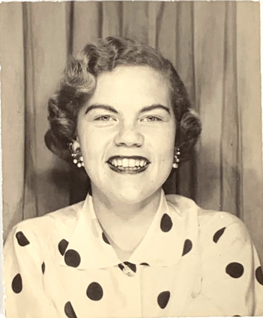 1950s vintage photo booth image of a young woman in a polkdot shirt and clip on earrings posing with a big smile.