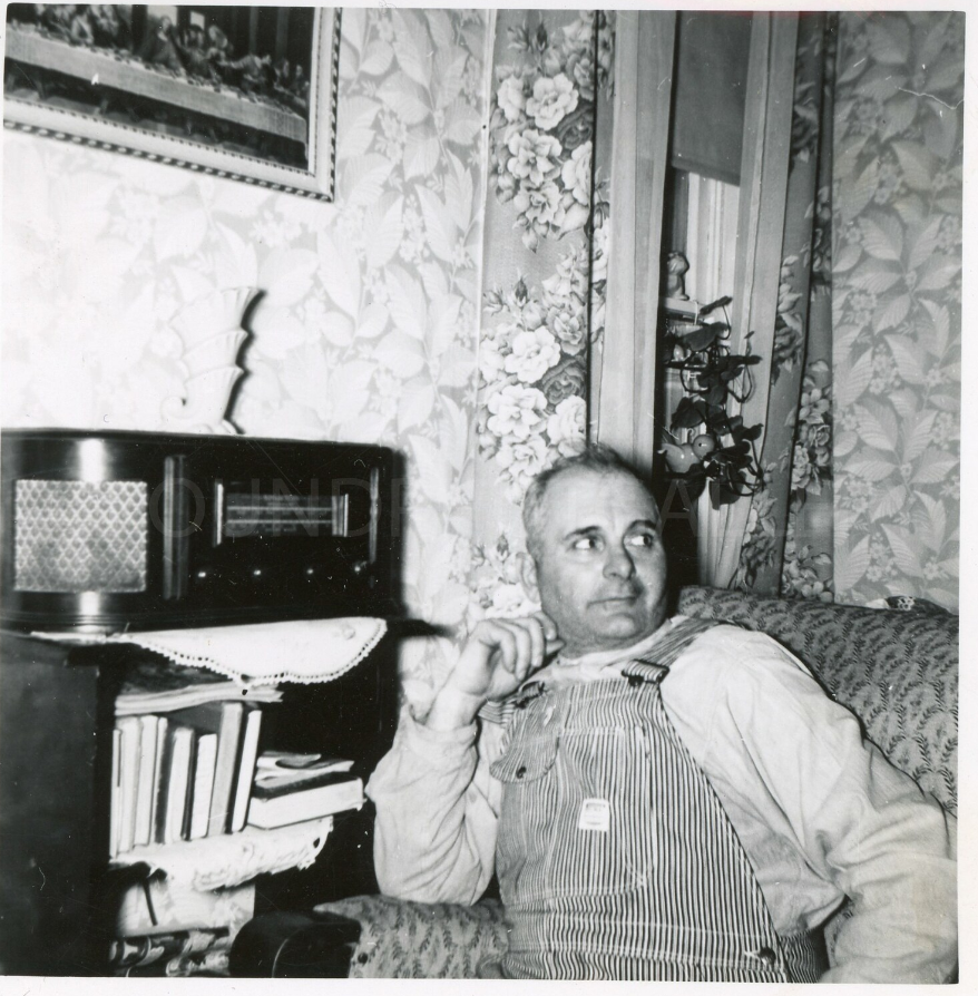 1940s vintage photo of a Burly Railroad Worker in Overalls Sitting in His Favorite Armchair By His Vintage Radio in a Cosy 1930’s Living Room