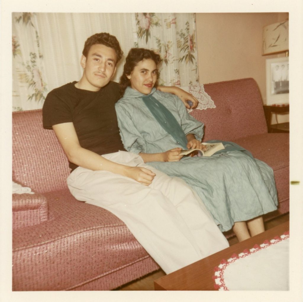 1960s vintage photo of a young couple sitting on a pink couch. The young woman is holding a book and in the background is a mid century lamp. 