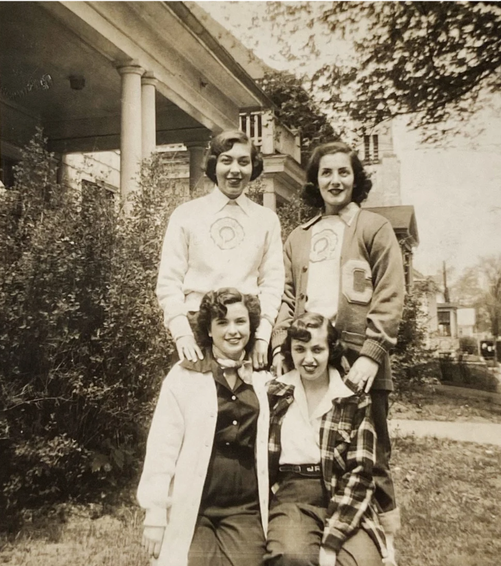 Early 1950s photo of 4 young college women in early 1950s fashions, posing together on the front lawn of their sorority house. One woman is wearing a letterman jacket. They have fantastic early 1950s hairstyles. 