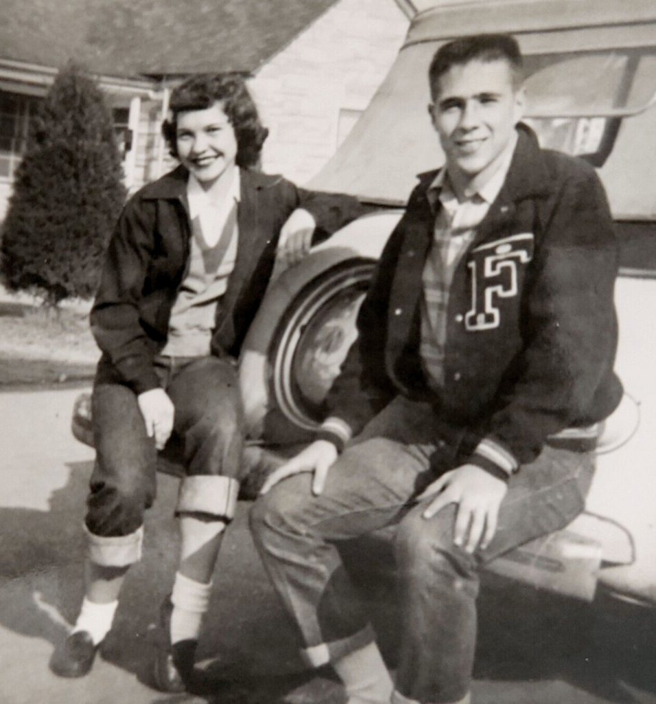 1950s vintage photo of two Teenagers sitting on the back of a car in 1950s fashions. The young man is wearing a Letterman Jacket. Photo is from 1954.