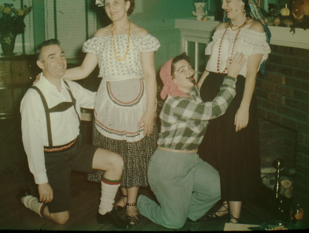 1950s vintage photo of a 1950s adult Halloween party featuring 3 women and a man. The man is in Lederhosen, while his partner is dressed in a dirndl skirt and peasant blouse. A woman is dressed up as a man in front of a woman in an eastern european outfit.