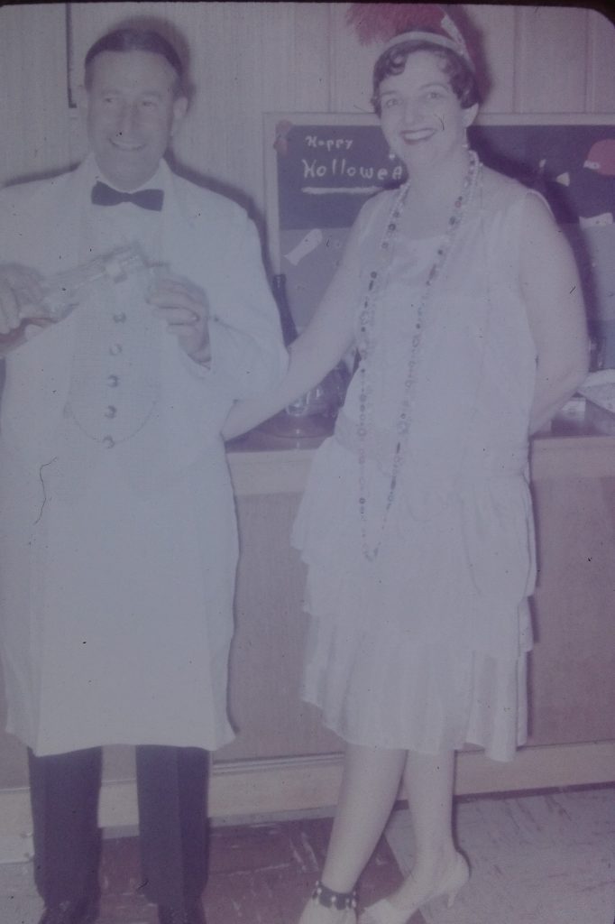 1960s vintage photo of a a man dressed as a bartender and a woman as a 1920s flapper pose for a picture at a 1967 party. 