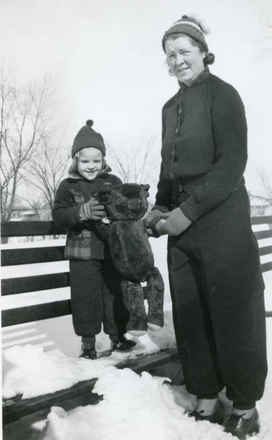 1930s vintage photo of a young girl in a snowsuit holding her teddy bear in teh snow with her mom in 1930s winter fashion stands beside her