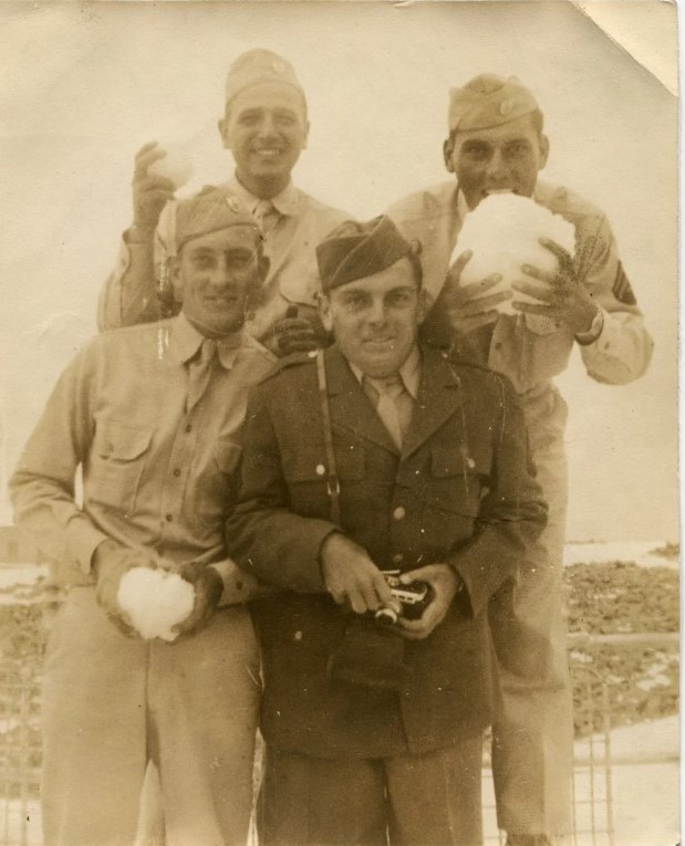 1940s vintage photo of 4 soldiers in uniform during WW2 posing outside in the snow holding giant snowballs and one man is holding a camera. 