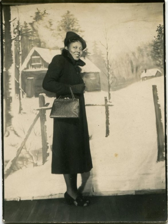 1940s vintage photo of a young Black woman posing in her winter coat with purse and hat in a photobooth with a winter landscape