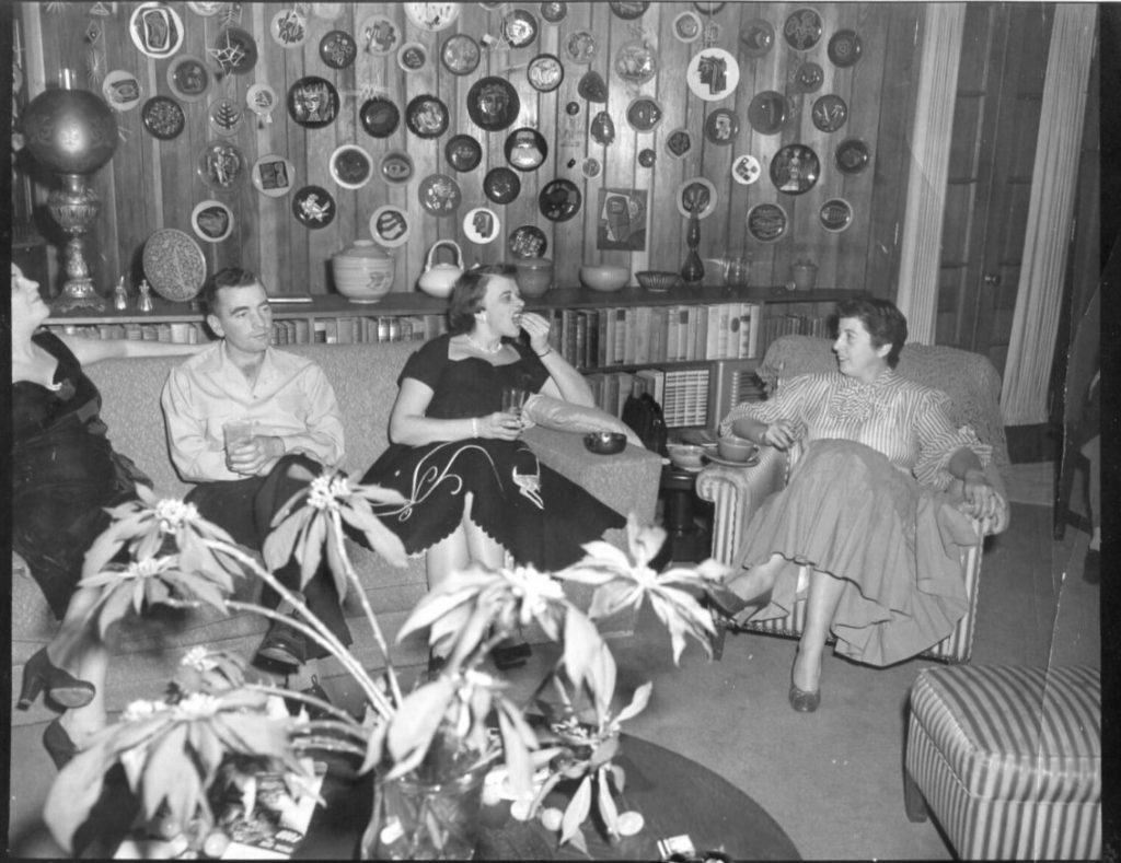 1950s vintage photo: 1950s Christmas party in a livingroom with a wall of plates and a well stocked bookshelf. The room has stylish 1950s women in 1950 fashions and one man. 