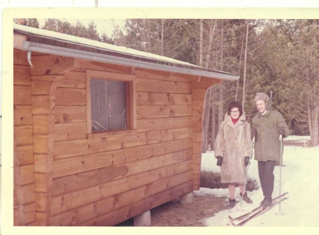 1960s photo of a husband and wife posing outside of their winter cabin as the man heads off to do some cross country skiing. 