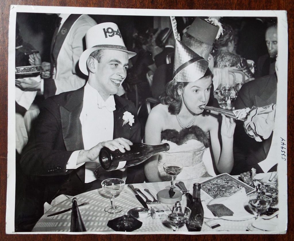 1939 going into 1940 vintage photo of a young couple celebrating New Year's Eve with champagne, horn blower and party hats. 