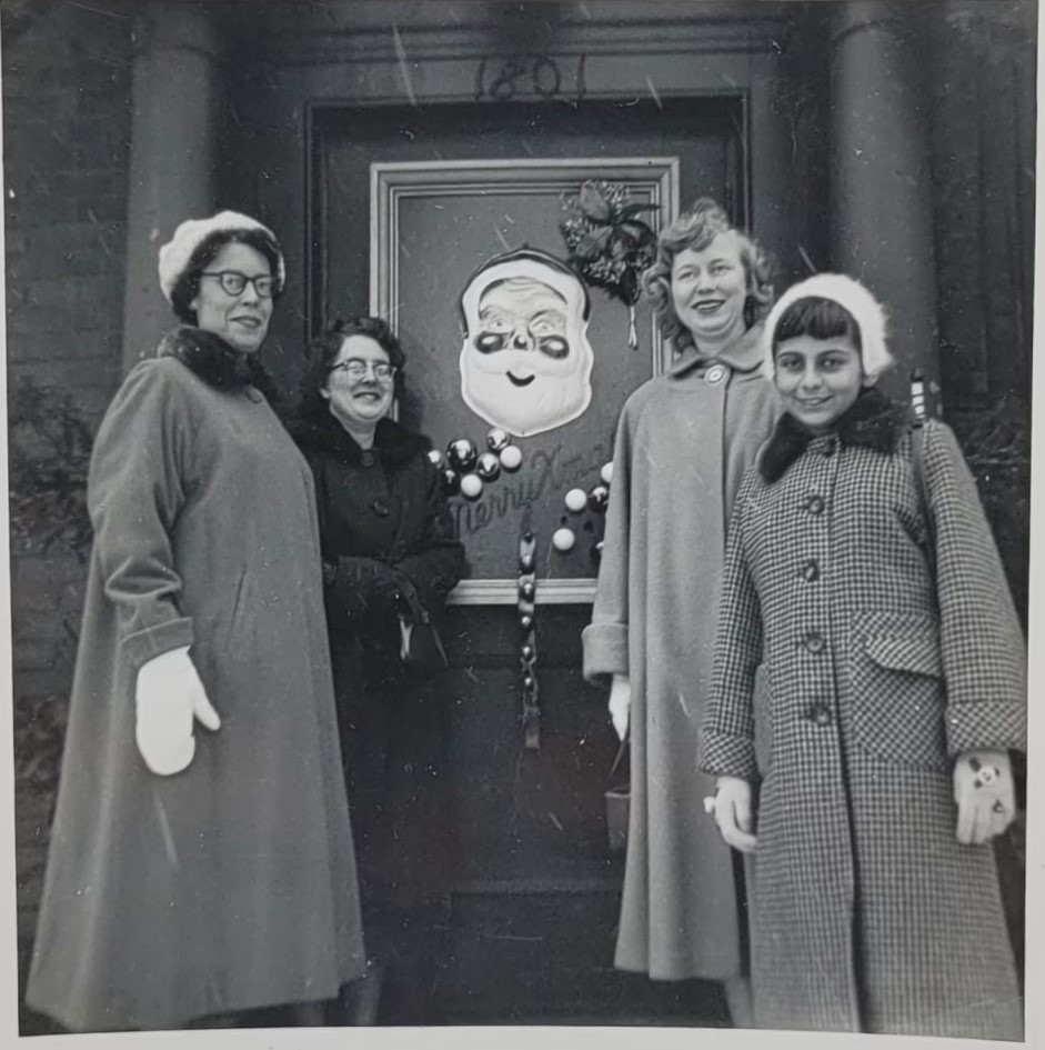 1950s vintage photo of 3 women & one young girl in 1950s coats posing with a mask of santa on the door of a building