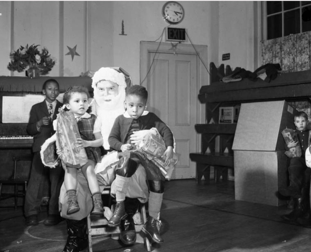 1950s vintage photo: Dec 19th, 1950 Santa pays a visit to a Black community centre. and has two little kids sitting on his lap with presents. 